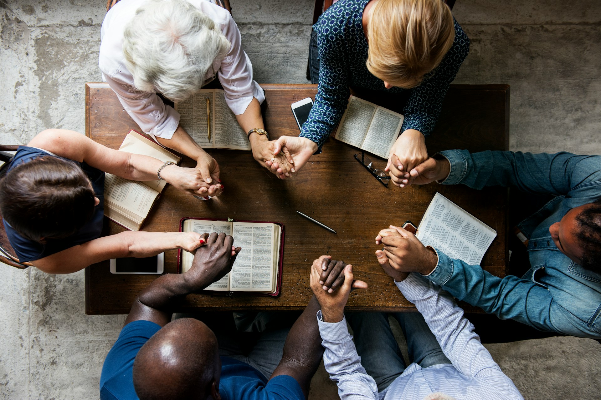 Overhead view of a group holding hands in prayer around a table with open Bibles